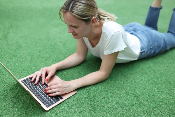 Mujer sonriente se encuentra en el césped de acuerdo con el ordenador portátil — Foto de Stock
