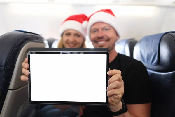 Leende man och kvinna i Tomten Claus hattar hålla tablett med tom vit skärm i flygplan stuga — Stockfoto