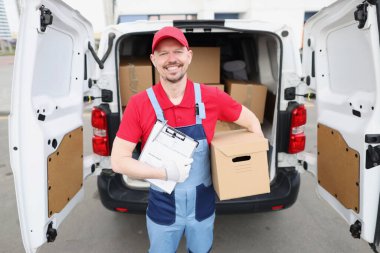 Adam kurye, aracın yanında teslimat belgeleri ve karton kutu tutuyor.