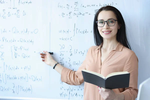 Wanita matematikawan dengan kacamata berdiri di papan tulis dengan rumus dan memegang buku terbuka — Stok Foto