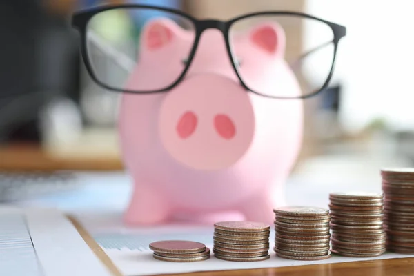 Pink piggy bank in glasses standing in front of piles of coins closeup — Stock Photo, Image