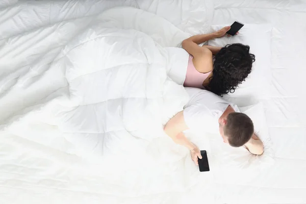 Homme et femme couchés dans la chambre à coucher sur le lit tenant les smartphones dans les mains et se détournant les uns des autres — Photo