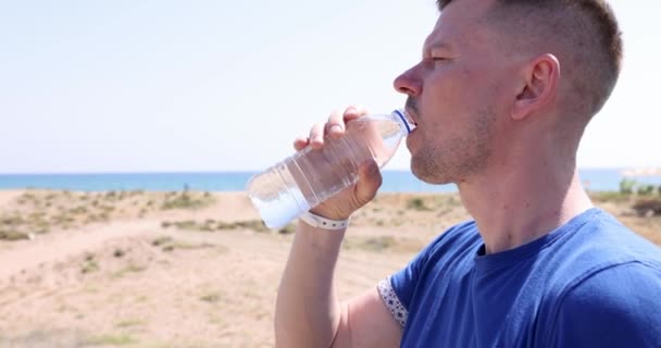 Jeune homme buvant de l'eau par la mer de bouteille en plastique en été film 4k — Video