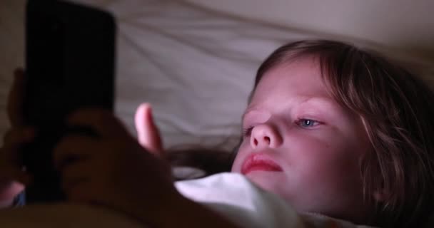 Liten flicka som ligger under filt och tittar på telefon skärm 4k film — Stockvideo