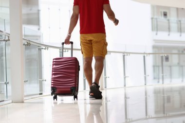 Burgundy bavulunu havaalanında yere çeken bir adam var.