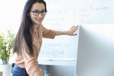 Kadın öğretmen elinde formüllerle karatahtanın önünde dizüstü bilgisayar ekranına bakıyor.