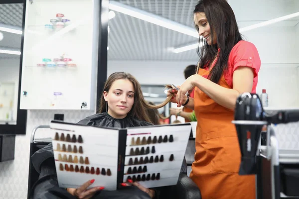 Giovane donna seduta in sedia parrucchiere e tenendo nel suo catalogo mani con tavolozza di colori per la tintura dei capelli — Foto Stock