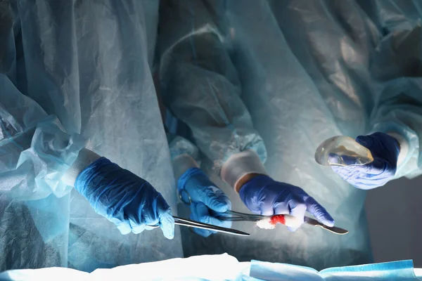 Doctors surgeons in protective coats holding instruments and breast implant in operating room closeup