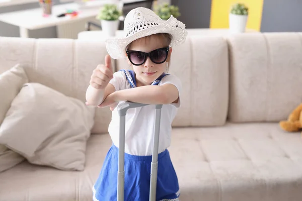 Menina em chapéu e óculos de sol fica com mala e segura os polegares para cima — Fotografia de Stock