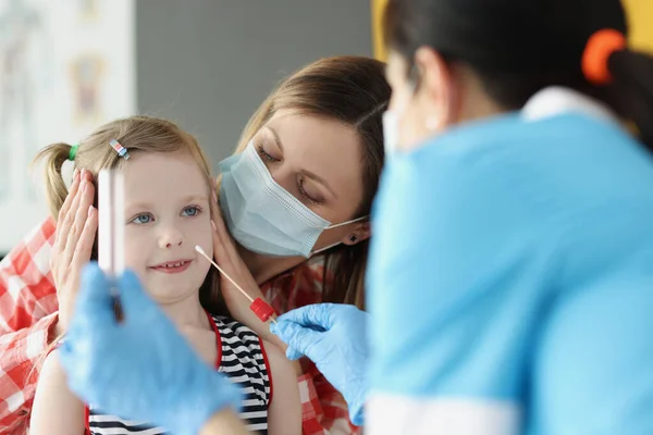 Dokter bertopeng pelindung membuat noda dengan kapas swab untuk gadis kecil di klinik — Stok Foto