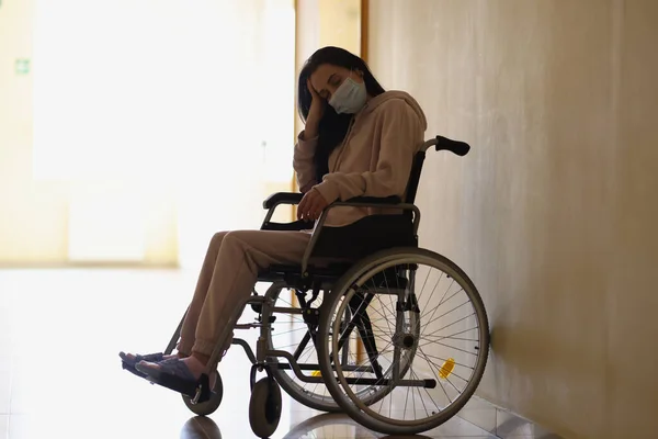 Молода жінка-інвалід у захисній медичній масці сидить у інвалідному візку в лікарняному коридорі — стокове фото