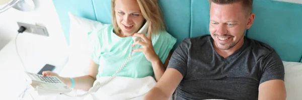 Muž a žena leží na posteli. Žena mluví po telefonu, muž přepíná tlačítka na dálkovém ovládání — Stock fotografie