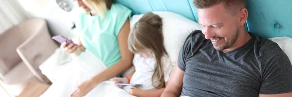 Os pais e a criança estão na cama, cada um segurando o smartphone em suas mãos — Fotografia de Stock