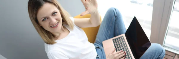 Młoda kobieta z laptopem siedząca w fotelu i machająca ręką — Zdjęcie stockowe