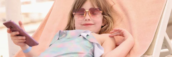 Pequeña chica sonriente en gafas de sol se encuentra en la tumbona con teléfono inteligente en sus manos — Foto de Stock