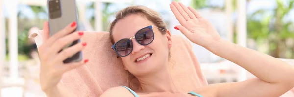 Retrato de una mujer sonriente tumbada en una tumbona en la playa y agitando su mano en un smartphone — Foto de Stock