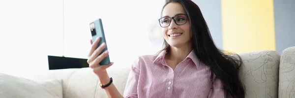 Mujer sonriente sentada en el sofá y mirando el teléfono inteligente — Foto de Stock