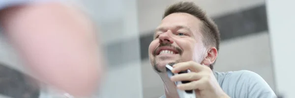 Человек бреет щетину с помощью машины перед зеркалом — стоковое фото