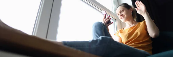 Jovem mulher sentada no peitoril da janela e acenando com a mão na tela do telefone celular — Fotografia de Stock