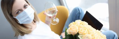 Koruyucu tıbbi maskeli bir kadın elinde bir bardak şarap ve elinde bir buket çiçek tutuyor.