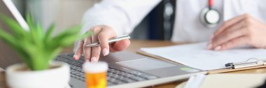 Doktorun elinde kalem var ve laptopta çalışıyor.