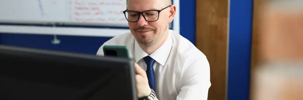 Νεαρός εξετάζει smartphone και οθόνη υπολογιστή στο χώρο εργασίας — Φωτογραφία Αρχείου