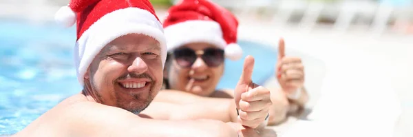 Homem e mulher em santa claus chapéus estão descansando no lado da piscina e mostrando o polegar para cima — Fotografia de Stock