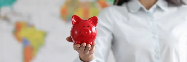 Young smiling woman holding red pig piggy bank on background of world map