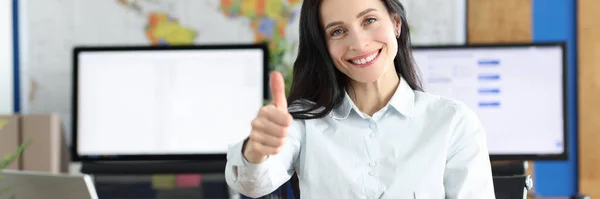 Joven mujer de negocios sonriente mostrando el gesto hacia arriba mientras está sentada en su mesa de trabajo en la oficina —  Fotos de Stock