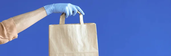 ブルーの背景に紙袋を保持ゴム手袋の宅配便の手 — ストック写真