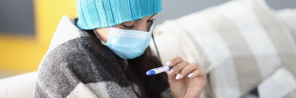 Woman in protective mask and warm hat sitting under covers and looking at thermometer