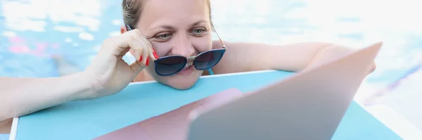 Mulher endireitando óculos de sol e olhando para a tela do laptop na piscina — Fotografia de Stock