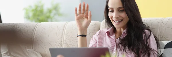 Young woman sitting on sofa and waving at laptop screen — Stock Photo, Image