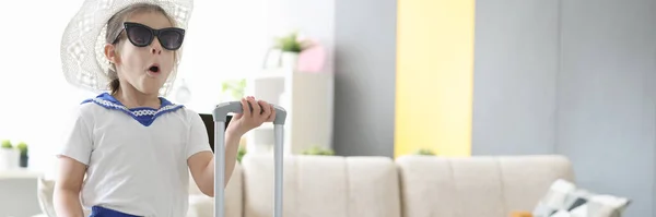 Sorpresa bambina in occhiali da sole e cappello in piedi con valigia in mano a casa — Foto Stock