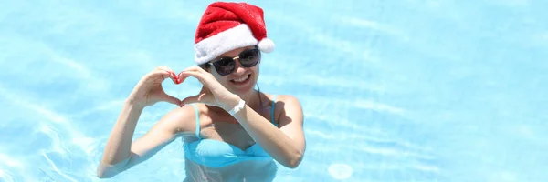 Усміхнена жінка в сонцезахисних окулярах і капелюсі Санта Клауса показує жест серця, стоячи в басейні — стокове фото