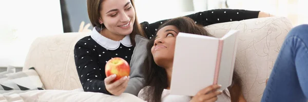 Duas jovens estão sentadas no sofá e lendo livro em casa — Fotografia de Stock