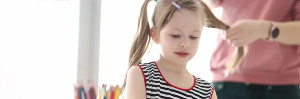 En liten flicka som ritar med pennor. Mor flätning barn hår — Stockfoto