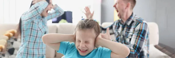 Little girl covering her ears with her hands against background of swearing parents at home — стоковое фото