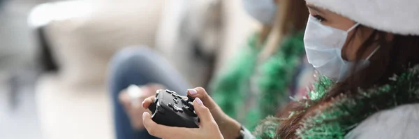 Dos novias están sentadas en el sofá con joysticks juego en sombreros de Santa Claus y máscaras médicas protectoras — Foto de Stock