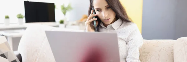 Mujer joven hablando por teléfono y trabajando en el ordenador portátil mientras está sentado en el sofá — Foto de Stock