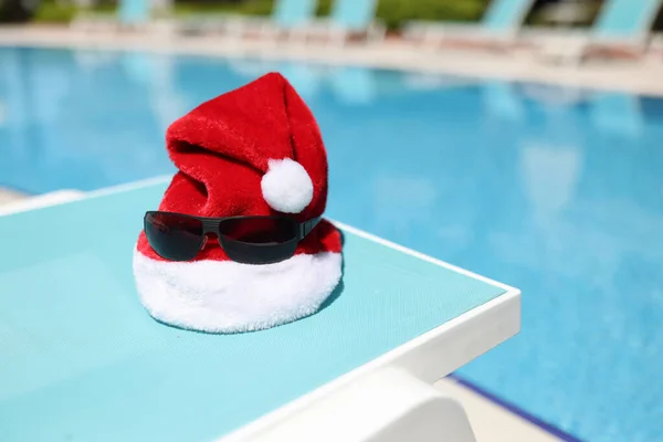 Chapeau santa claus rouge et lunettes de soleil couché sur la chaise longue sur le rivage de la piscine gros plan — Photo