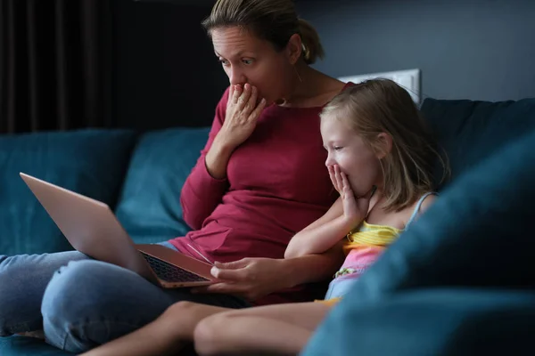 Sorprendidos mamá y su hija están sentados en el sofá y mirando a la pantalla del ordenador portátil —  Fotos de Stock