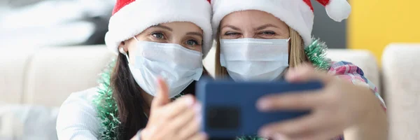 Duas mulheres em máscaras médicas protetoras e chapéus de Papai Noel tirar selfie no smartphone — Fotografia de Stock