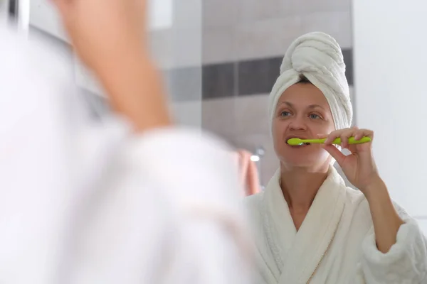 Giovane donna in accappatoio e con asciugamano sulla testa e lavarsi i denti davanti allo specchio in bagno — Foto Stock