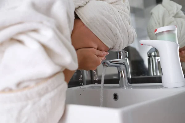 Jovem com toalha na cabeça lavagem rosto sob torneira no banheiro — Fotografia de Stock