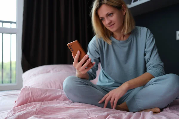 Молода жінка в піжамі сидить у ліжку з мобільним телефоном в руці — стокове фото