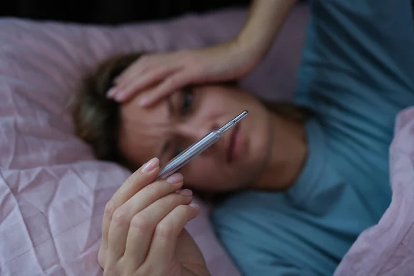 Sick woman lying in bed and holding thermometer with high temperature closeup