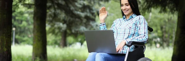 Young disabled woman in wheelchair waving hand at laptop screen in park