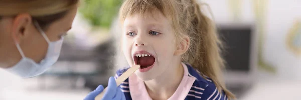 Öronhals yngolog läkare i skyddande medicinsk mask undersöka halsen på liten flicka med trä spatel — Stockfoto