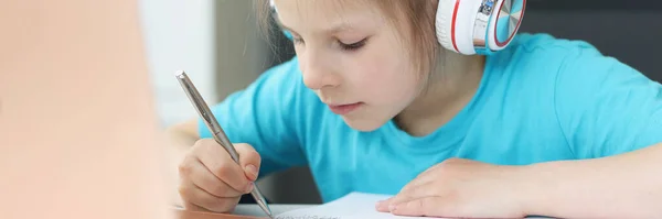 Маленькая девочка в наушниках сидит за столом перед ноутбуком и рисует — стоковое фото